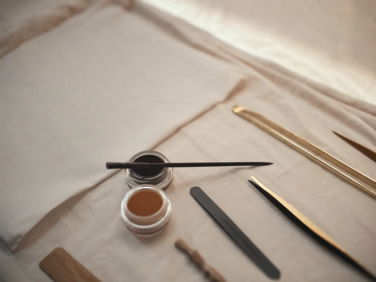 Professional brow tools — brush, tint pot and slanted tweezers on cream linen